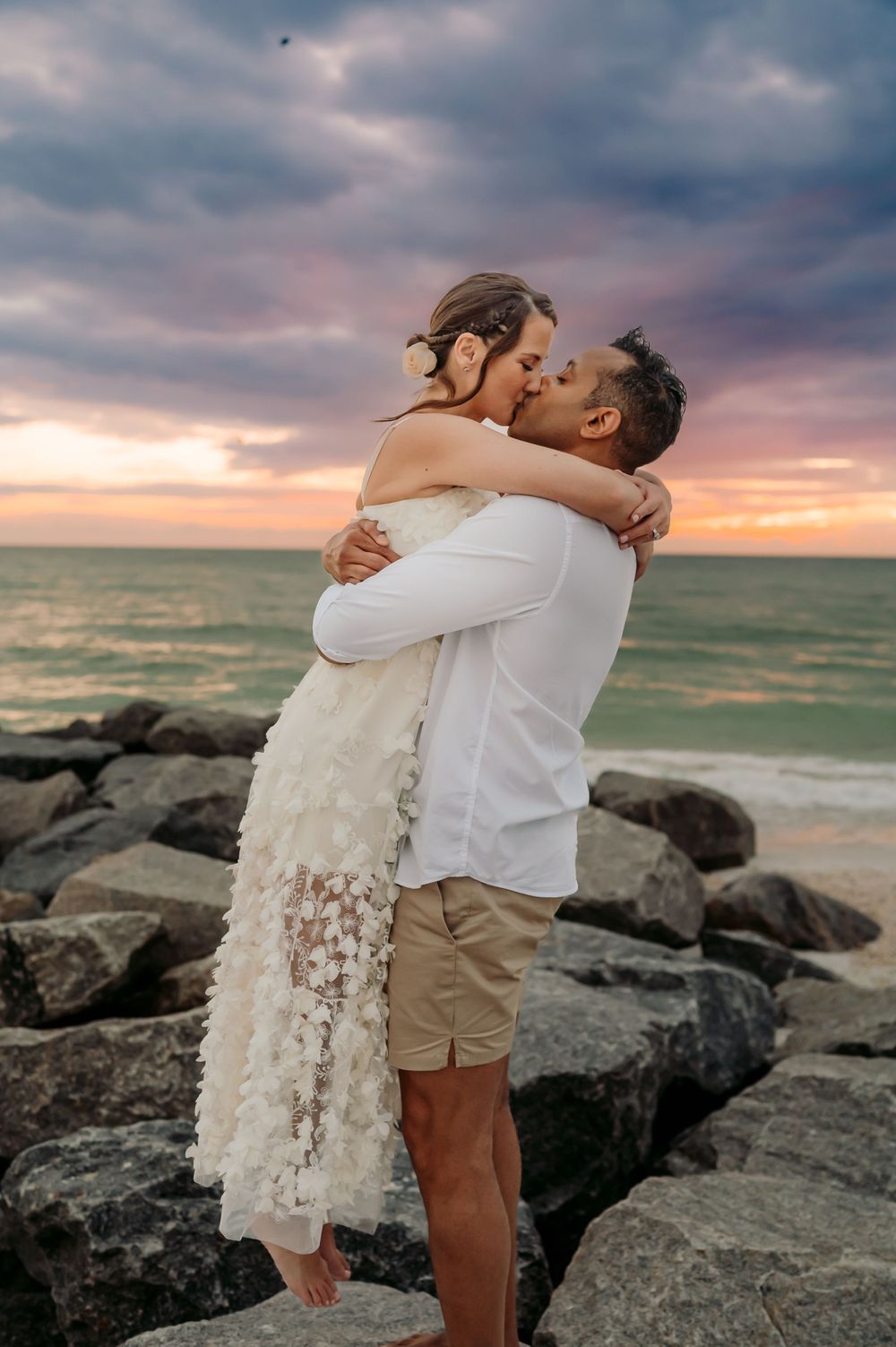 A newly-wed couple embraces and kiss in front of a spectacular, colorful sunset.