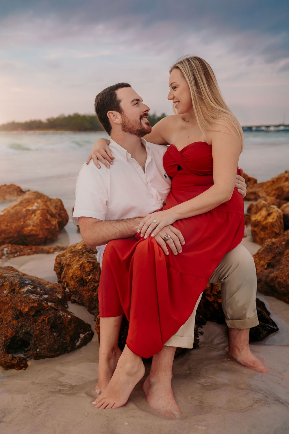 Man in white shirt and dress pants and woman in red dress sit on large rocks at the beach, looking at one another.