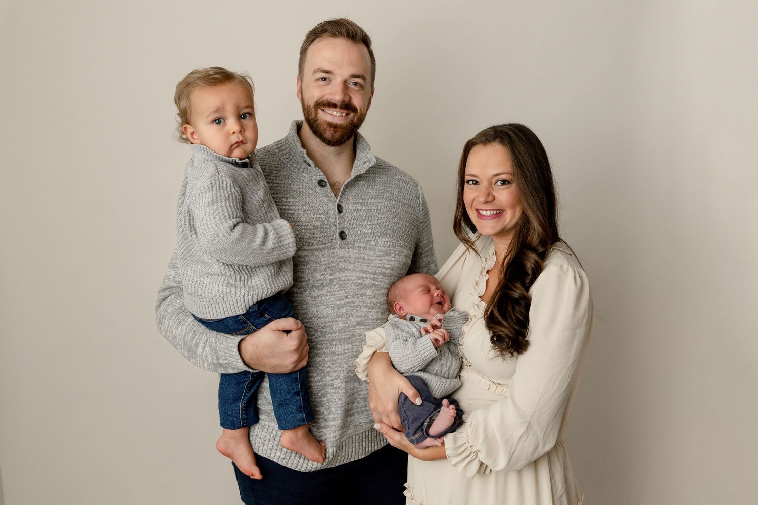 A family portrait against a neutral background shows parents with their toddler and newborn baby wearing coordinated grey clothing.