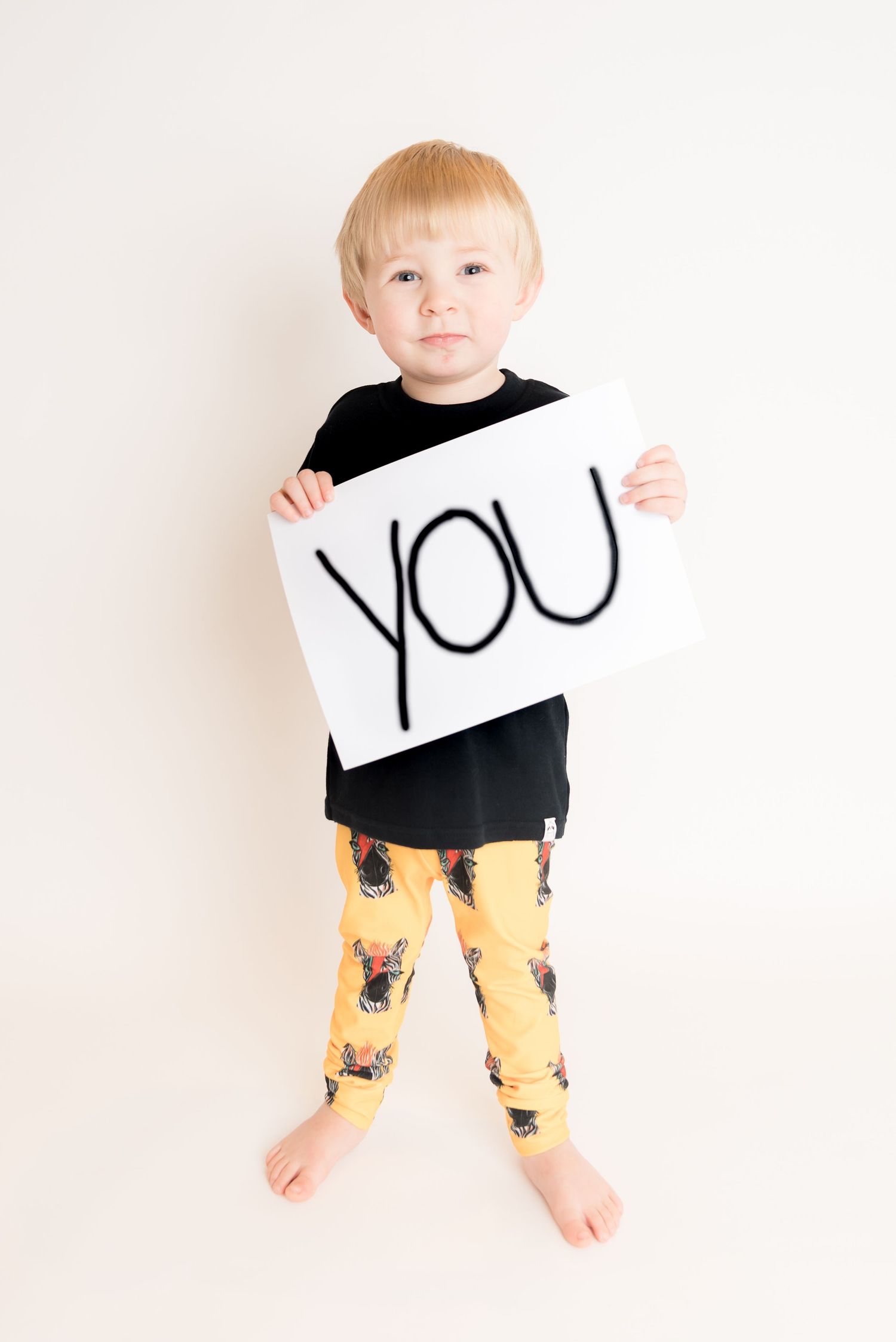 Small person wearing black shirt and yellow patterned pants holding a 'YOU' sign.