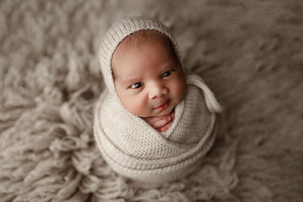 Newborn wrapped in cozy white knit blanket and bonnet on textured background.