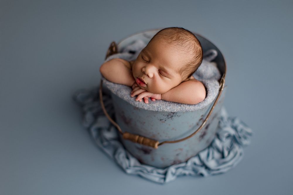 Sleeping newborn nestled in cozy gray bucket prop against blue backdrop.