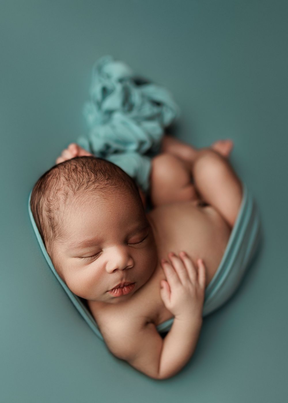 Sleeping infant nestled in teal fabric wrap during professional newborn photoshoot.
