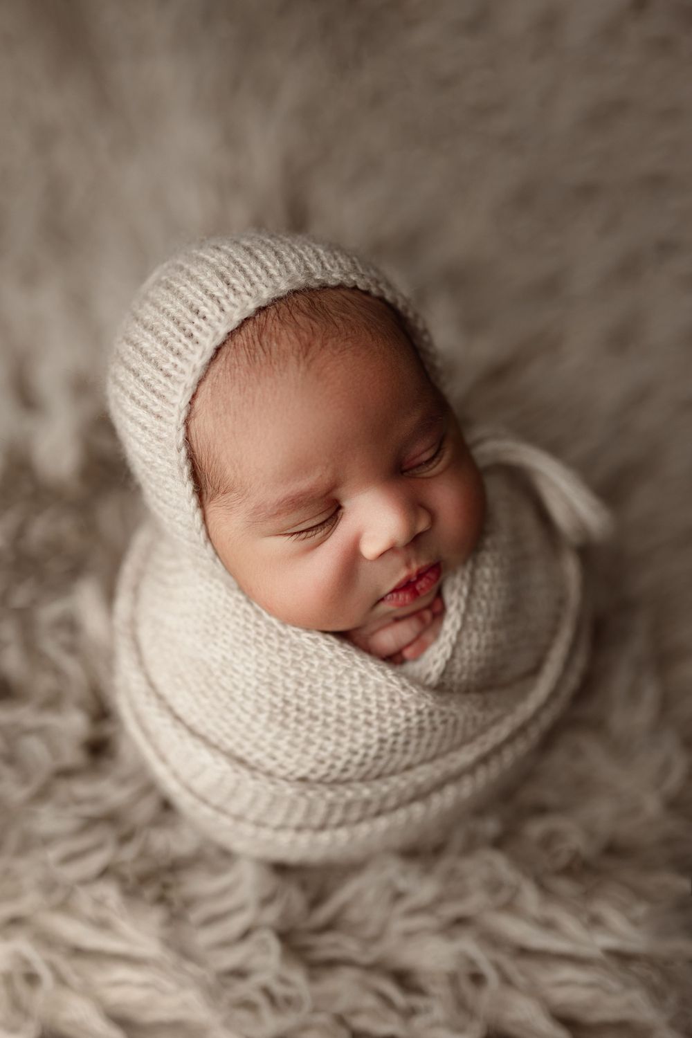 Sleeping newborn in cream knit wrap and bonnet photographed from above on fuzzy backdrop.