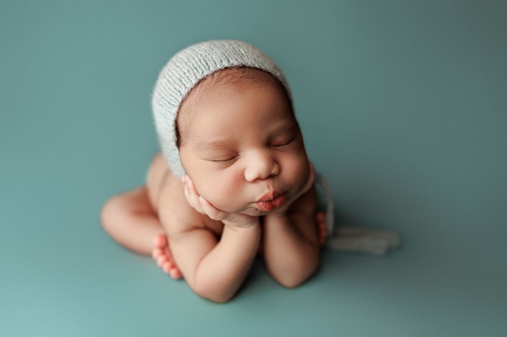 Sleeping newborn wearing white knit bonnet posed on teal background.