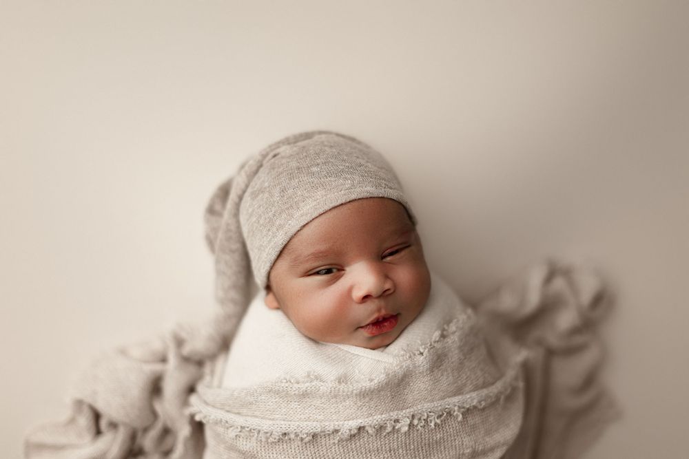 Newborn in gray and white wraps with knit cap against neutral background.