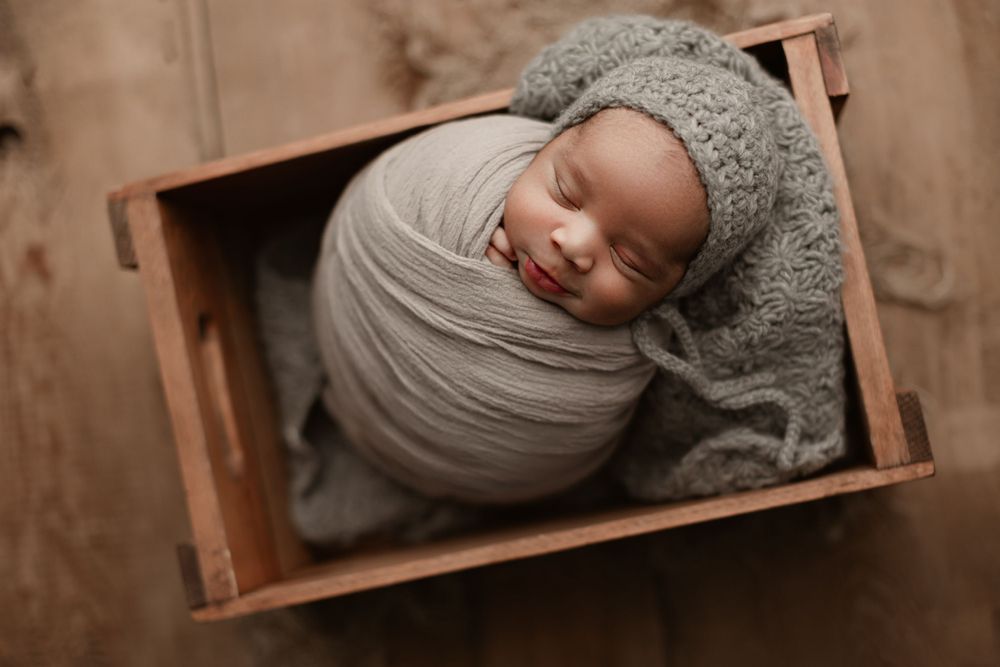 Peaceful newborn swaddled in gray wrap sleeping in wooden box.