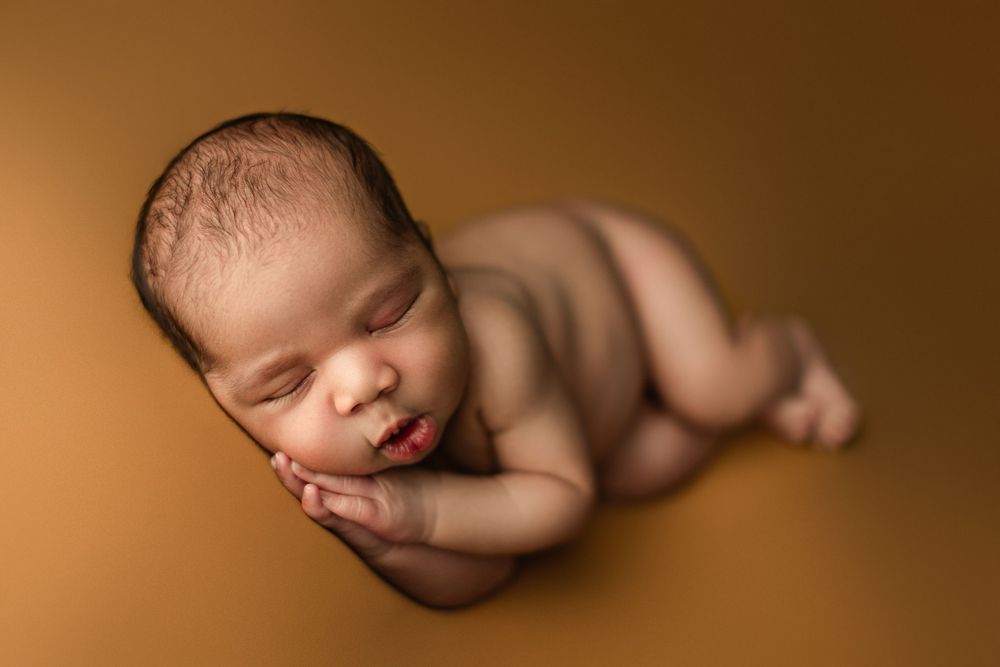 Sleeping newborn posed on warm brown backdrop in artistic portrait style.