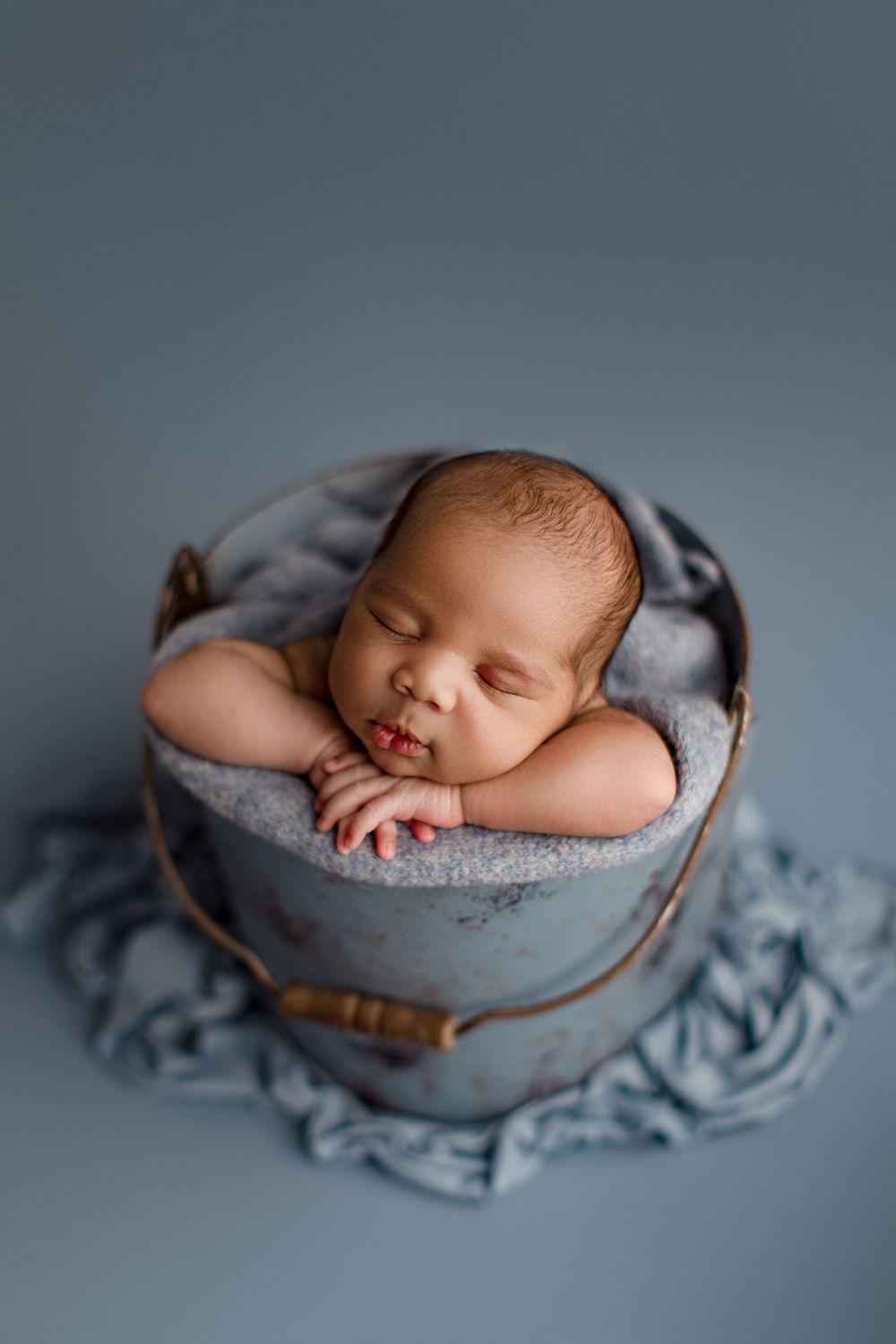 Peaceful newborn sleeping in vintage bucket prop with textured gray fabric.