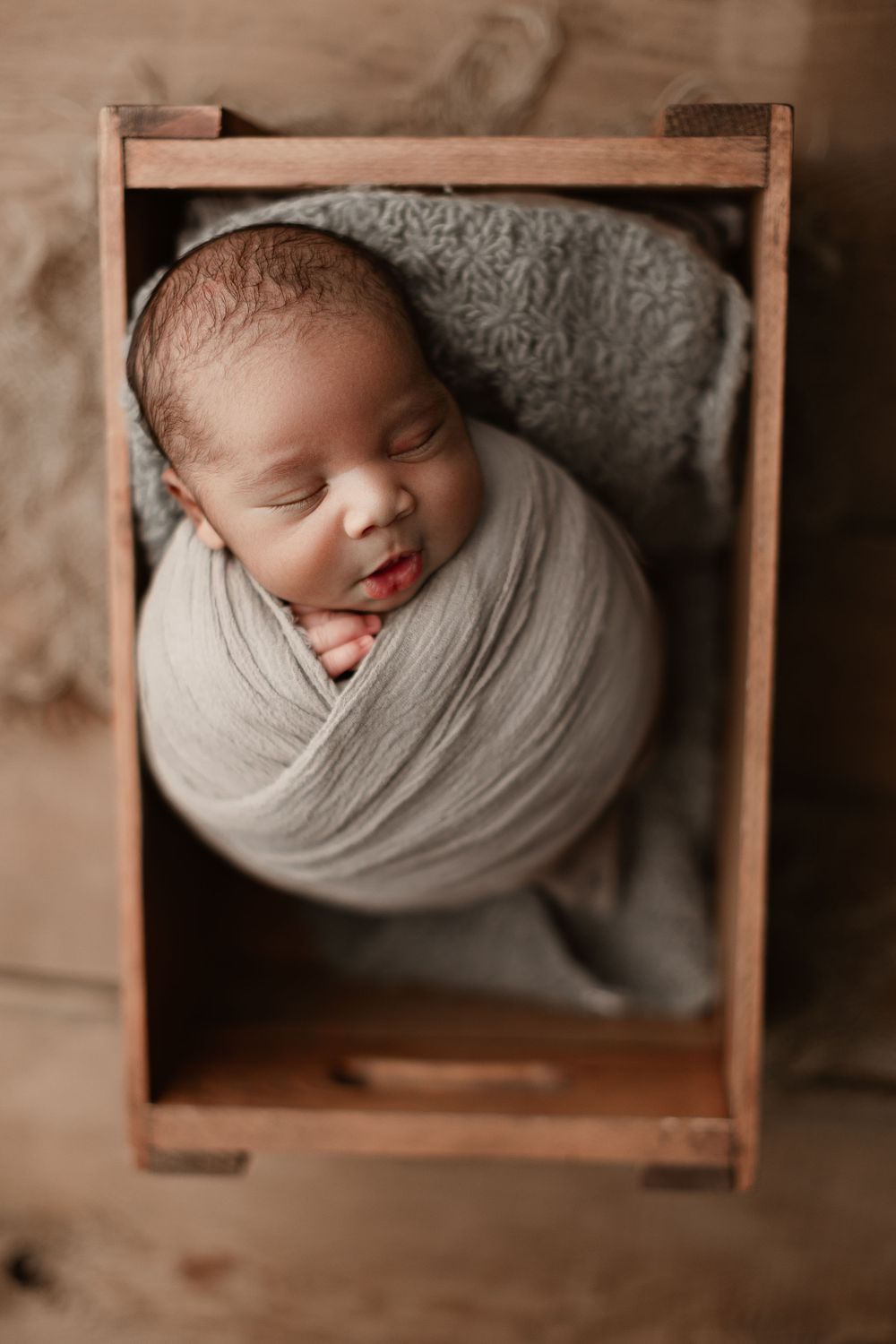 Smiling newborn wrapped in soft gray fabric resting in vintage wooden crate.