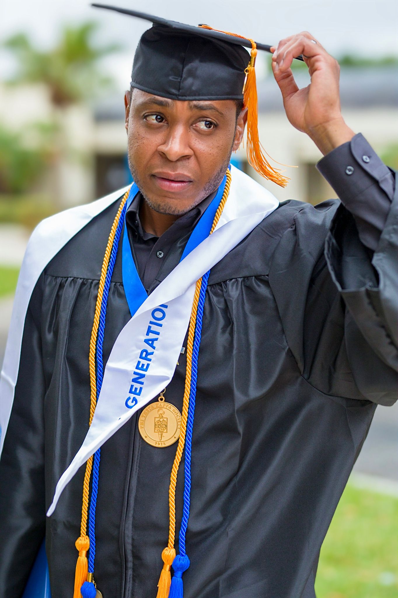 Graduate in cap and gown with blue and gold honor cords adjusting mortarboard.