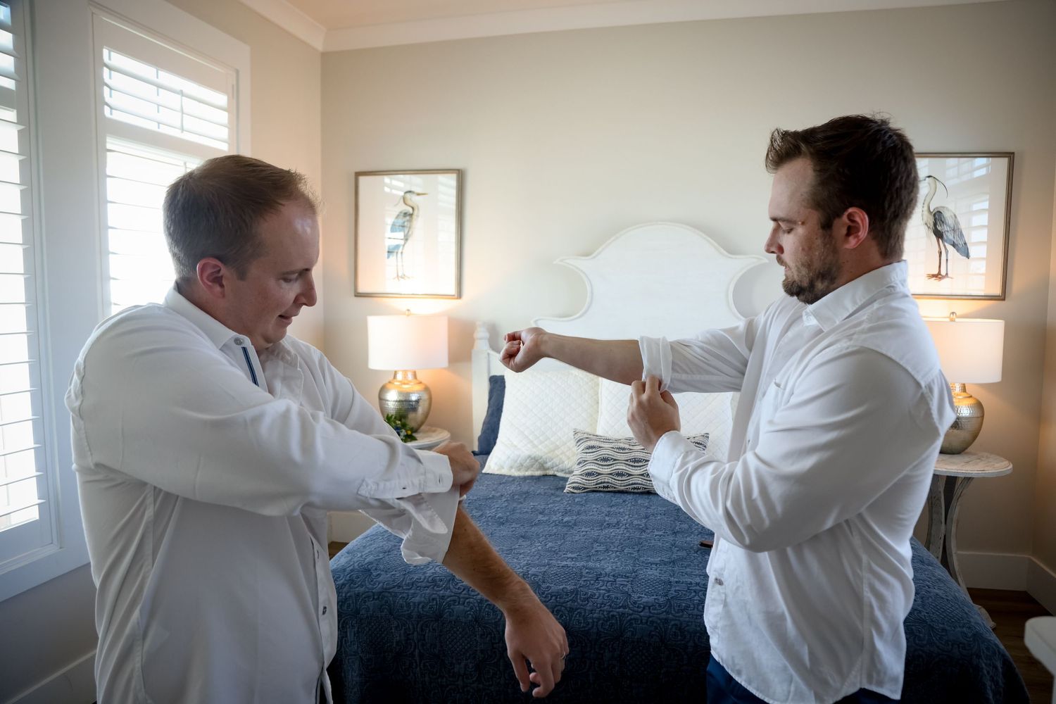 Two men in white dress shirts helping each other with cufflinks in a bedroom.