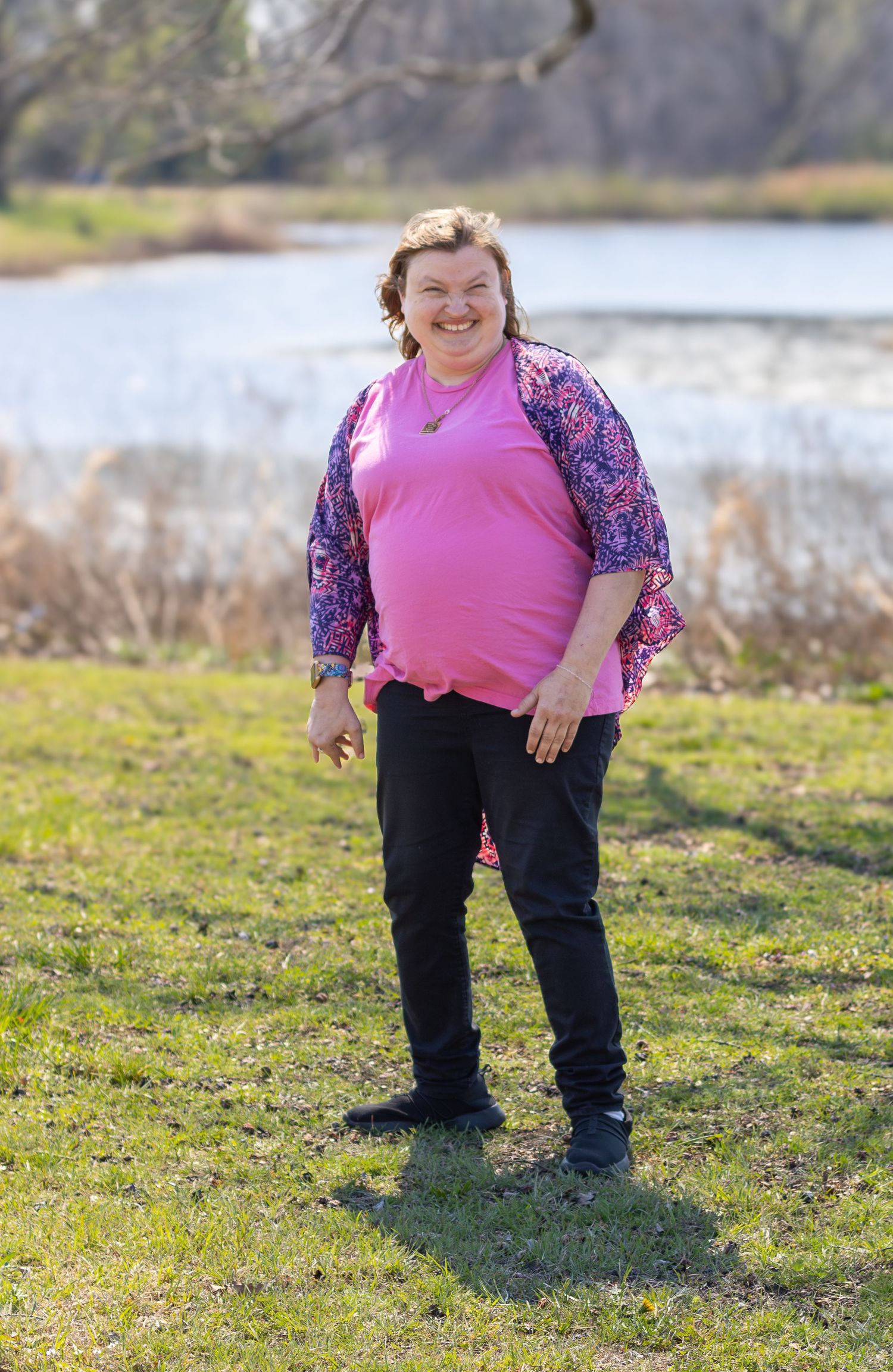 Person in pink top and floral sleeves posing on grass near a lake on a sunny day.