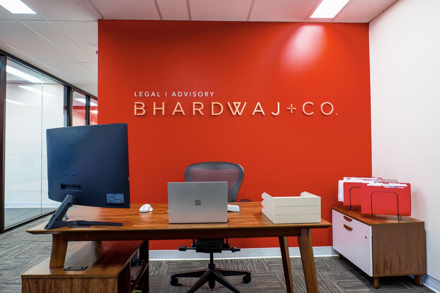 Modern office desk setup with Bhardwaj+Co legal branding on a bright orange wall.