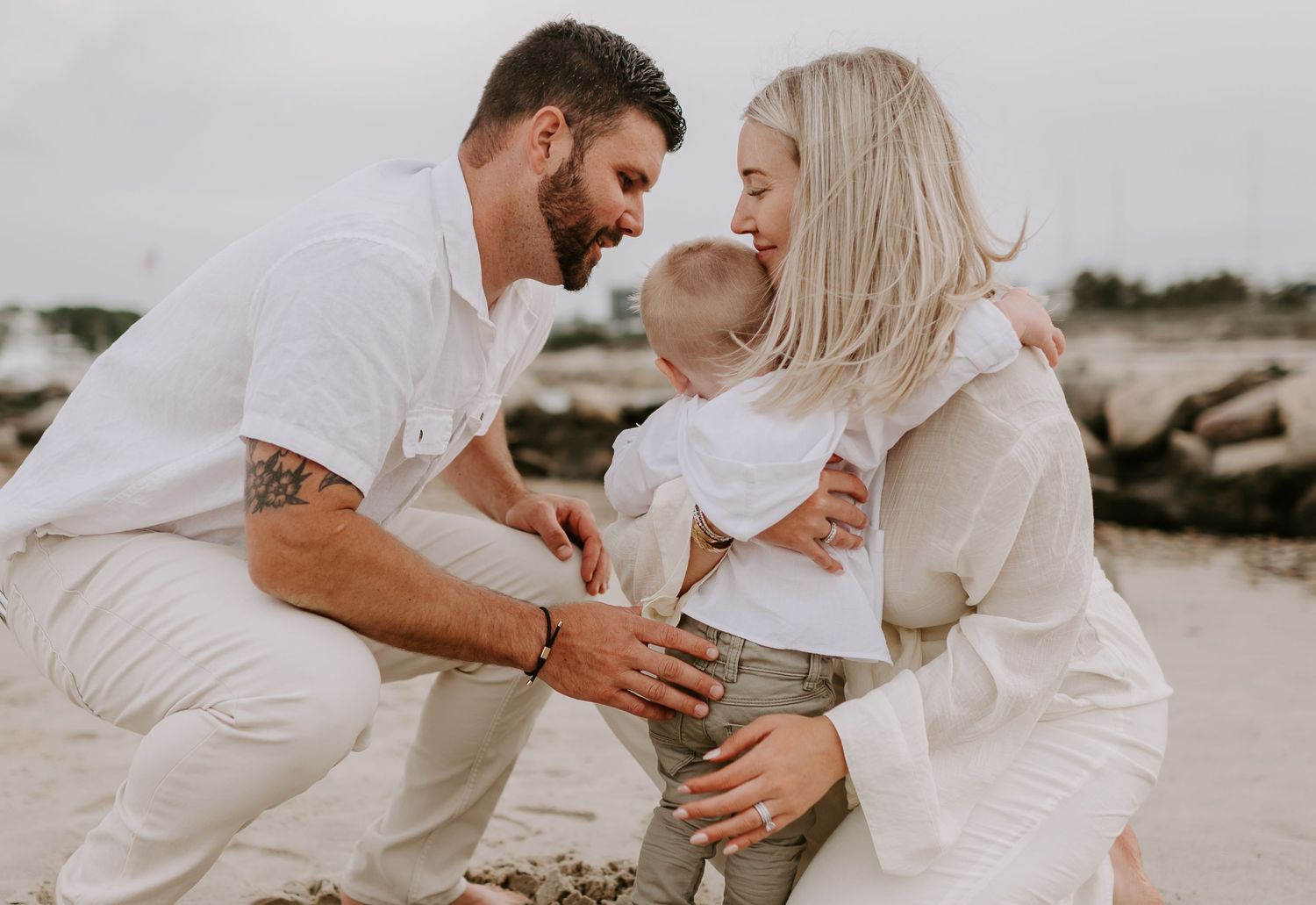 A couple dressed in white embraces on a sandy beach.