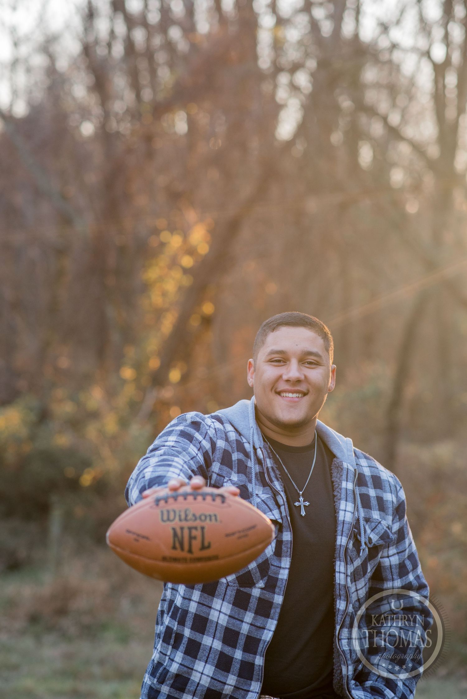 A person in plaid holds up an NFL Wilson football while posing in front of autumn trees.