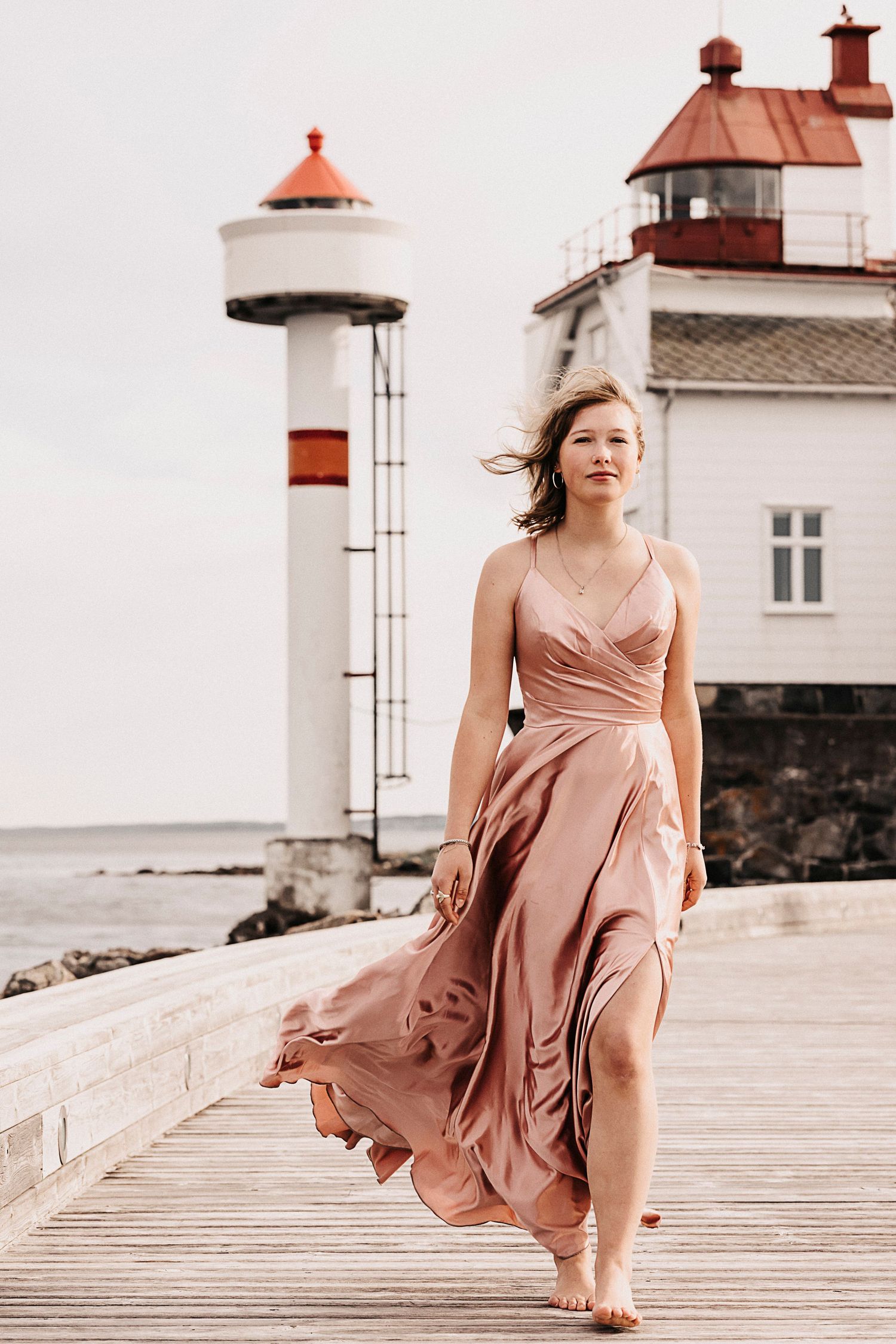 A flowing blush pink dress catches the wind on a wooden boardwalk near a lighthouse and white building by the sea.