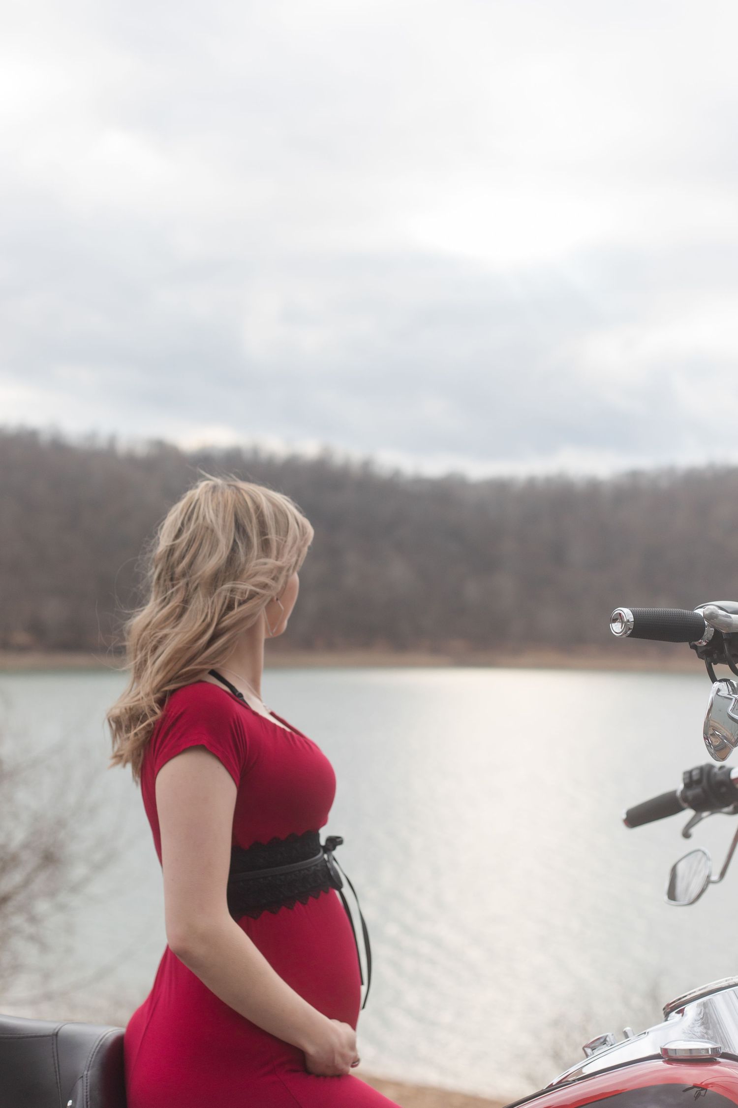A person in a red dress stands beside a motorcycle overlooking a winter lake landscape.