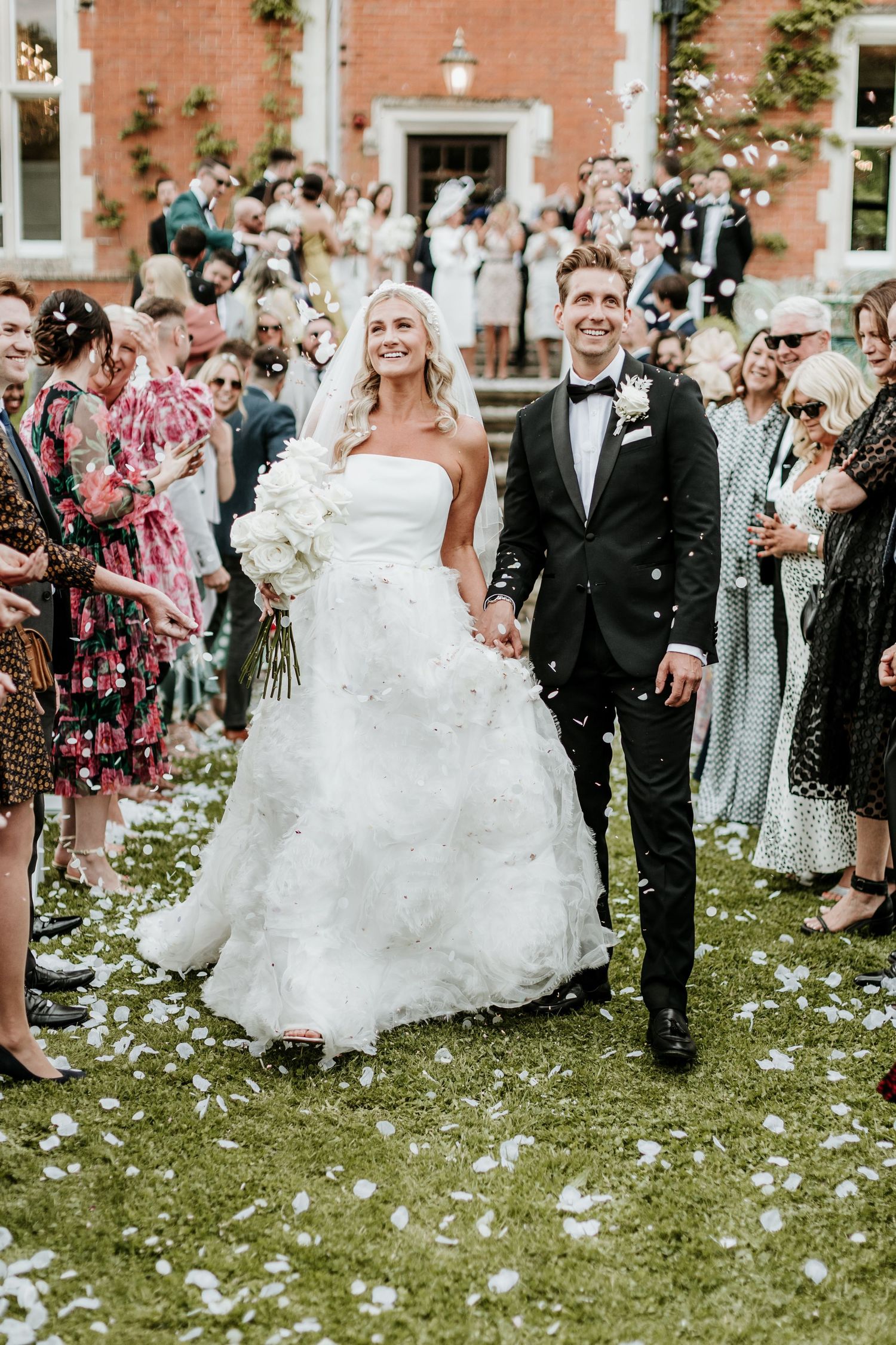 Newlyweds walk through a crowd of wedding guests throwing flower petals outside a brick building.
