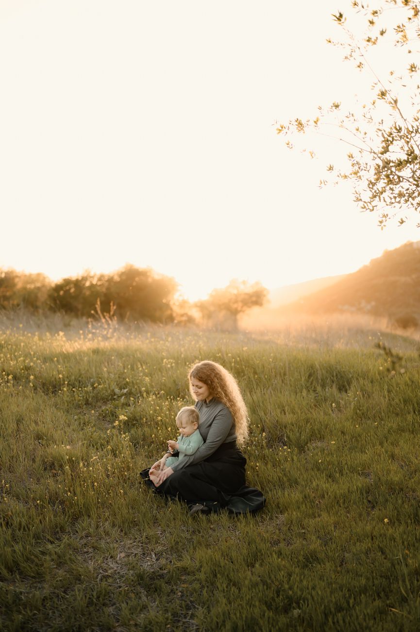 Motherhood portrait session by Escondido Photographer, of woman sitting in flower field with baby at sunset