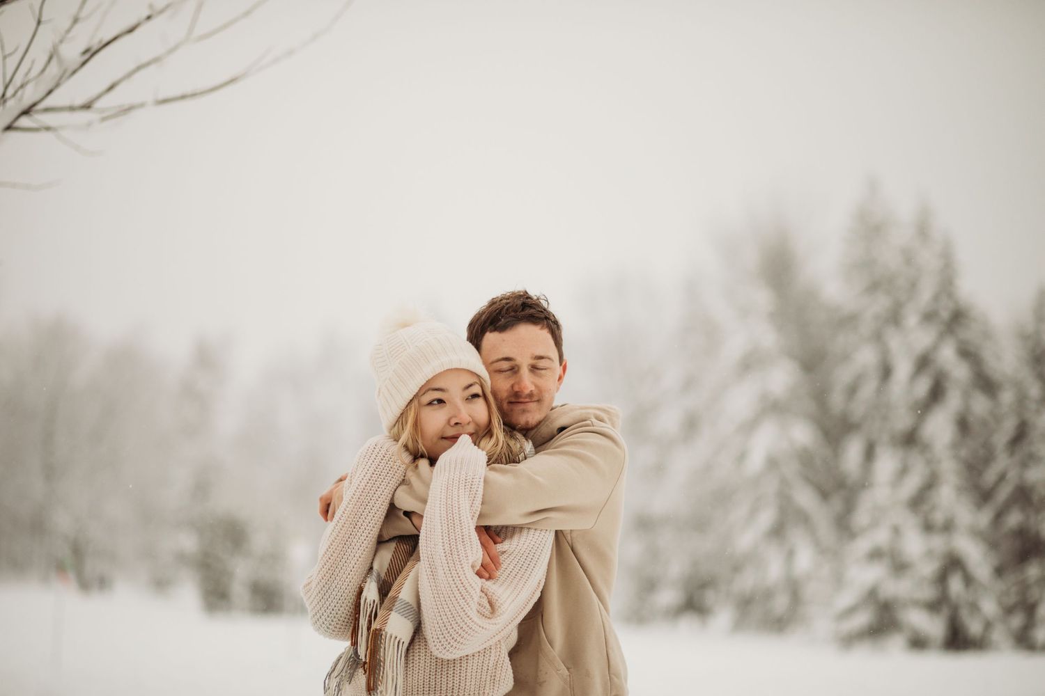 A romantic winter photoshoot with a couple laughing while wrapped in warm sweaters against a snowy forest.