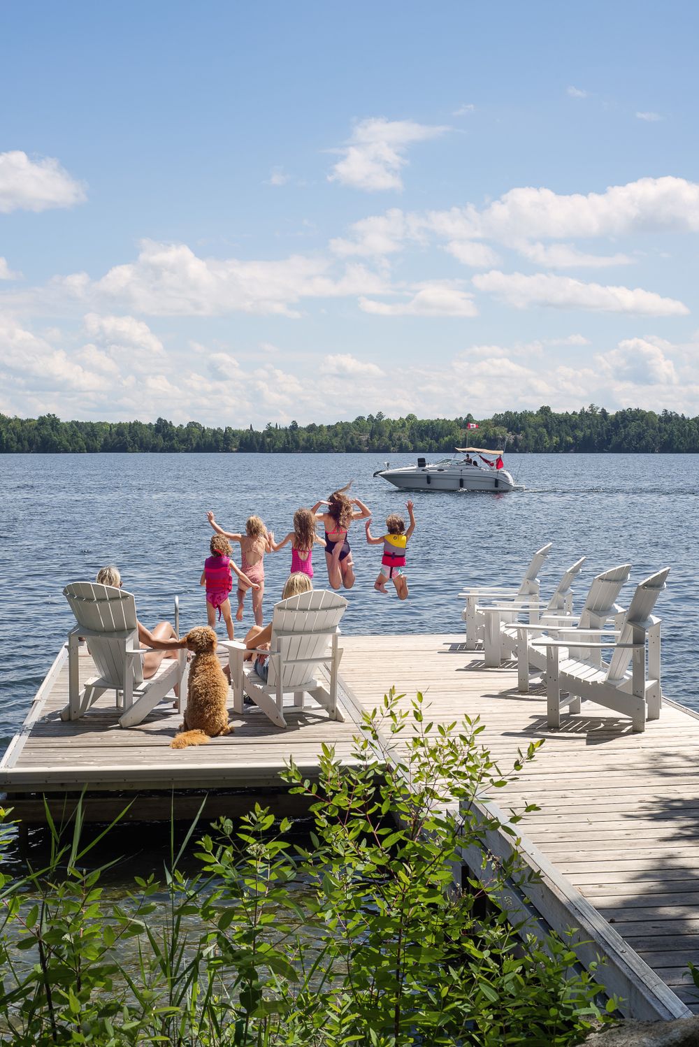 Summer lake scene with white Adirondack chairs on wooden dock and motorboat in background.