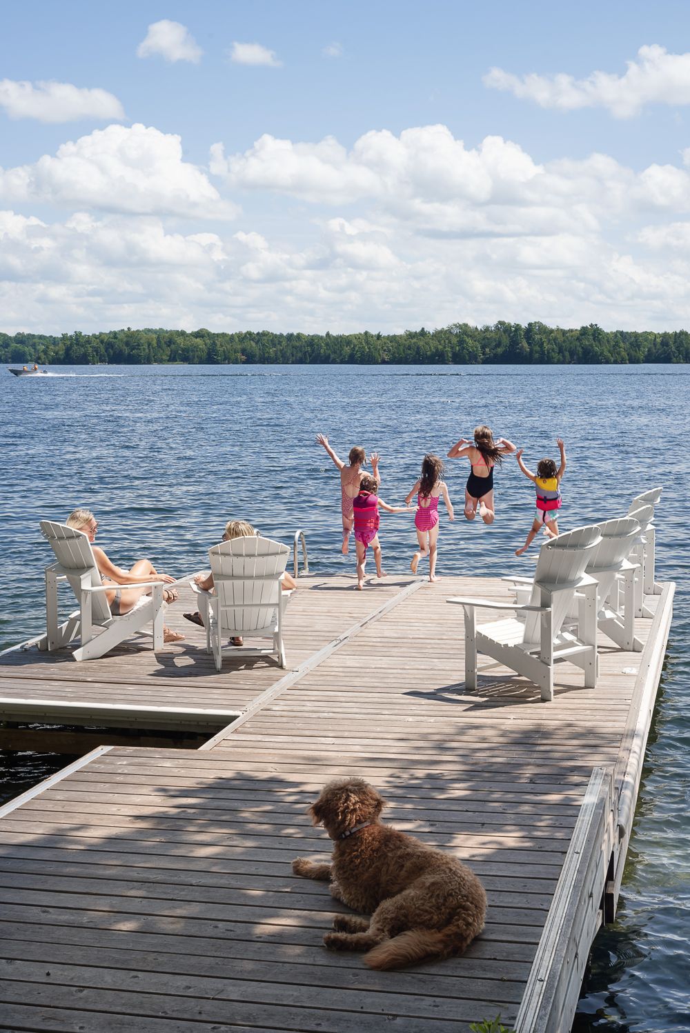People enjoying a summer day on a wooden dock with Adirondack chairs and a dog while others jump into the lake.