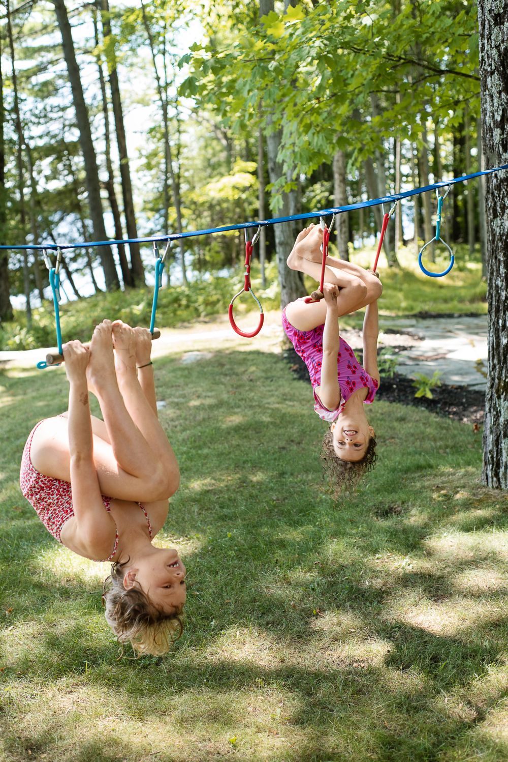 Two children hanging upside down on gymnastic rings in a backyard with trees and lake in background.