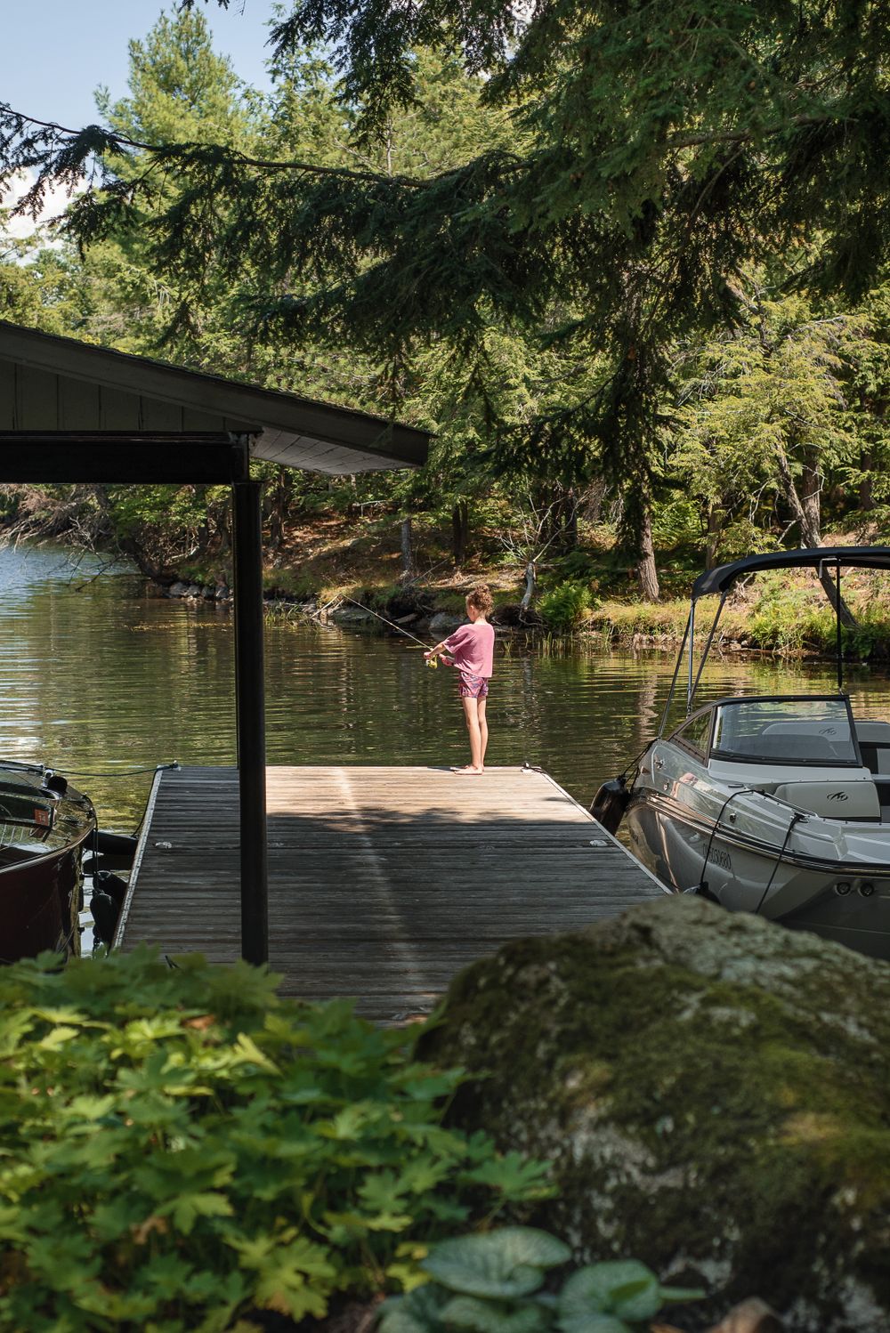 Person standing on a lakeside dock with boats and dense forest in the background.