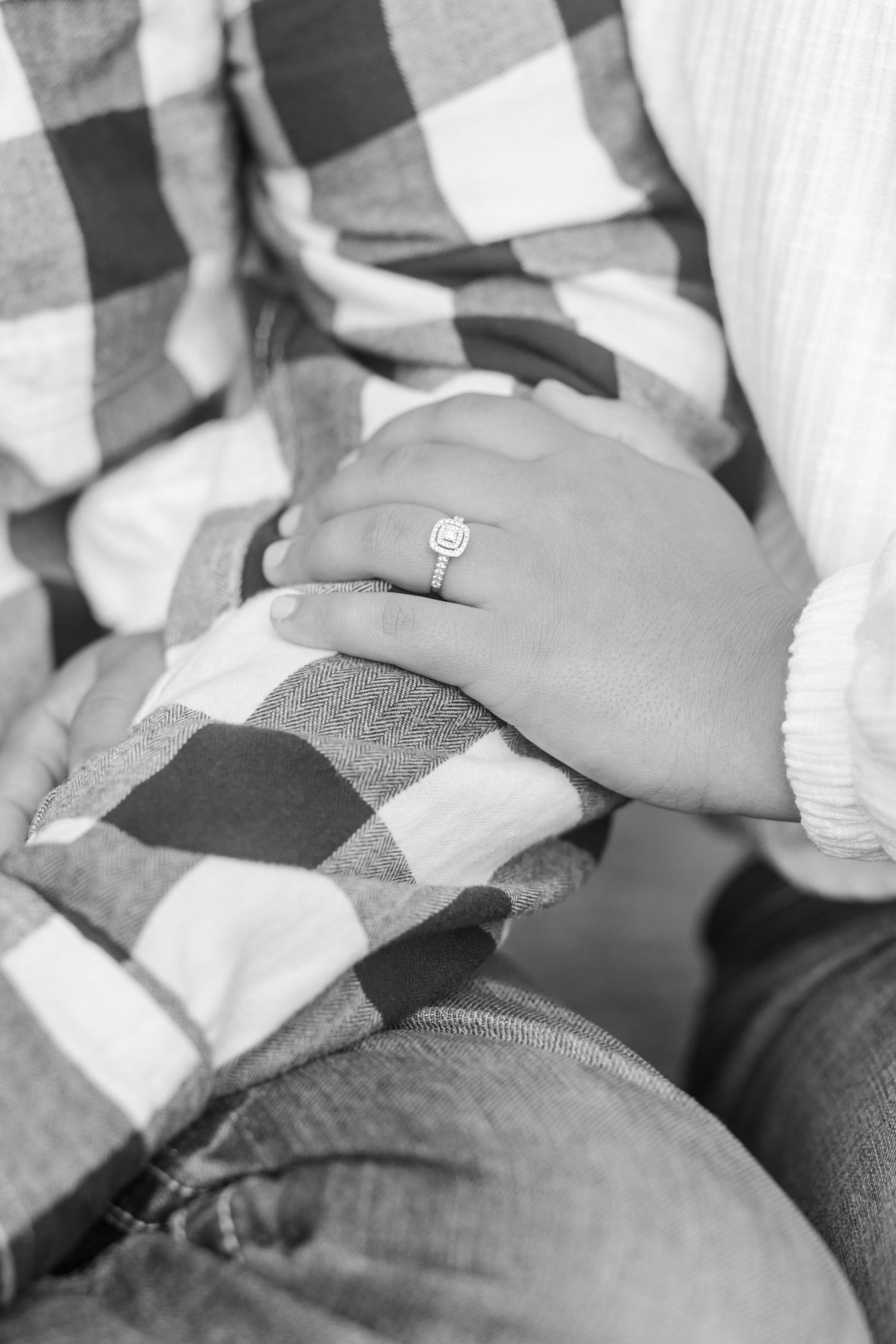 Black and white photo of hands with engagement ring resting on buffalo plaid blanket.