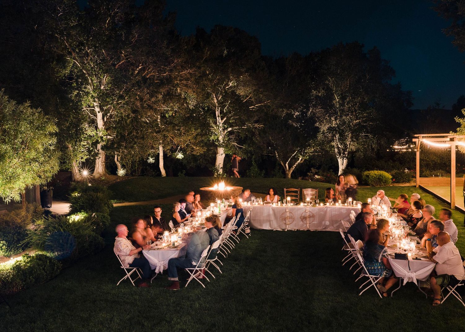 Outdoor evening dinner party with string lights illuminating tables under trees.