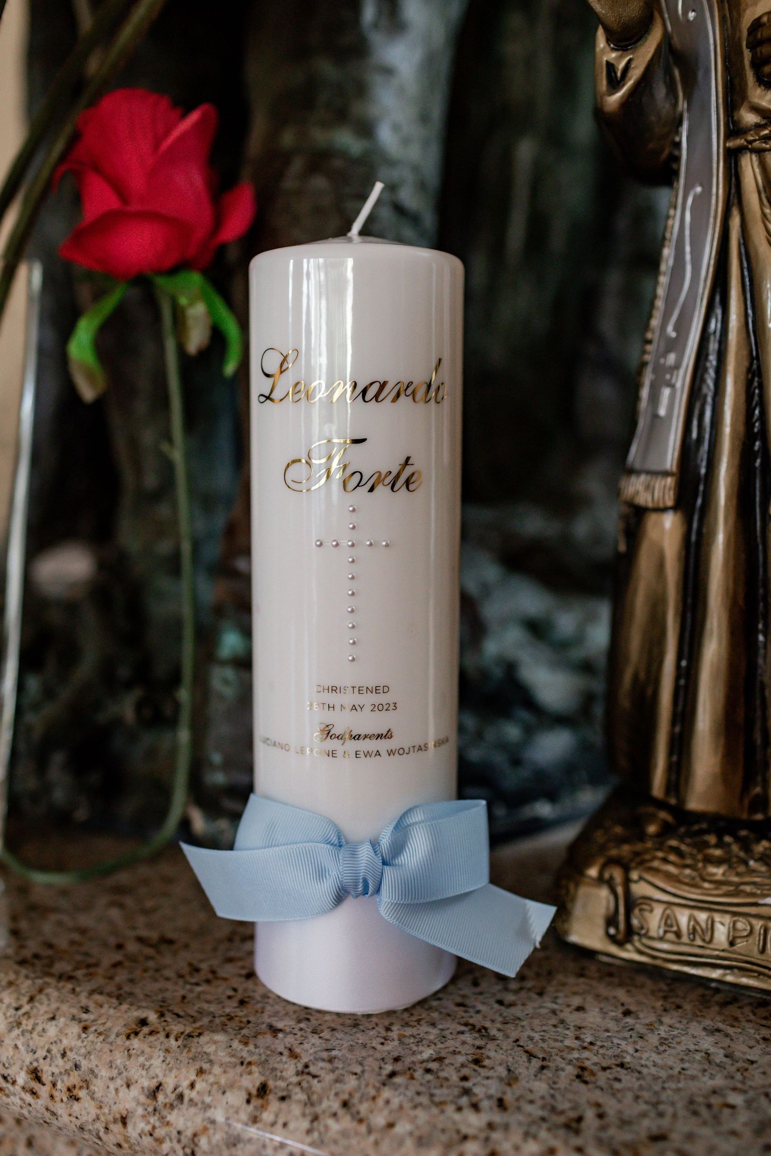 Decorative white candle with gold text and light blue ribbon next to a red rose.