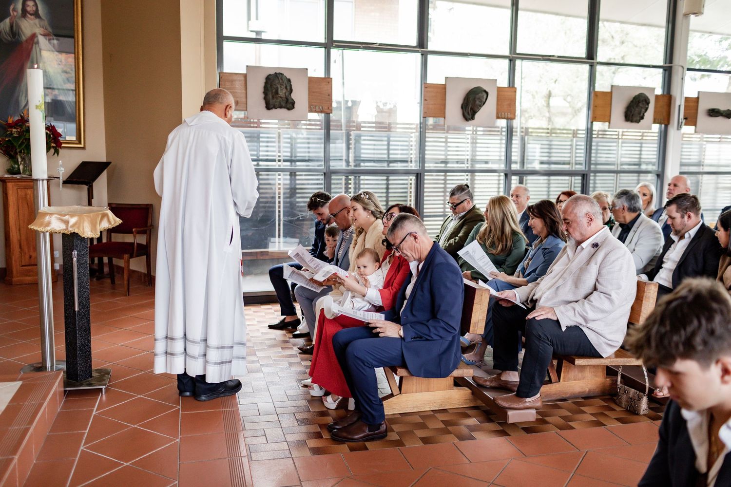 A clergy member in white robes leads a church service with attendees seated in rows on a tiled floor.