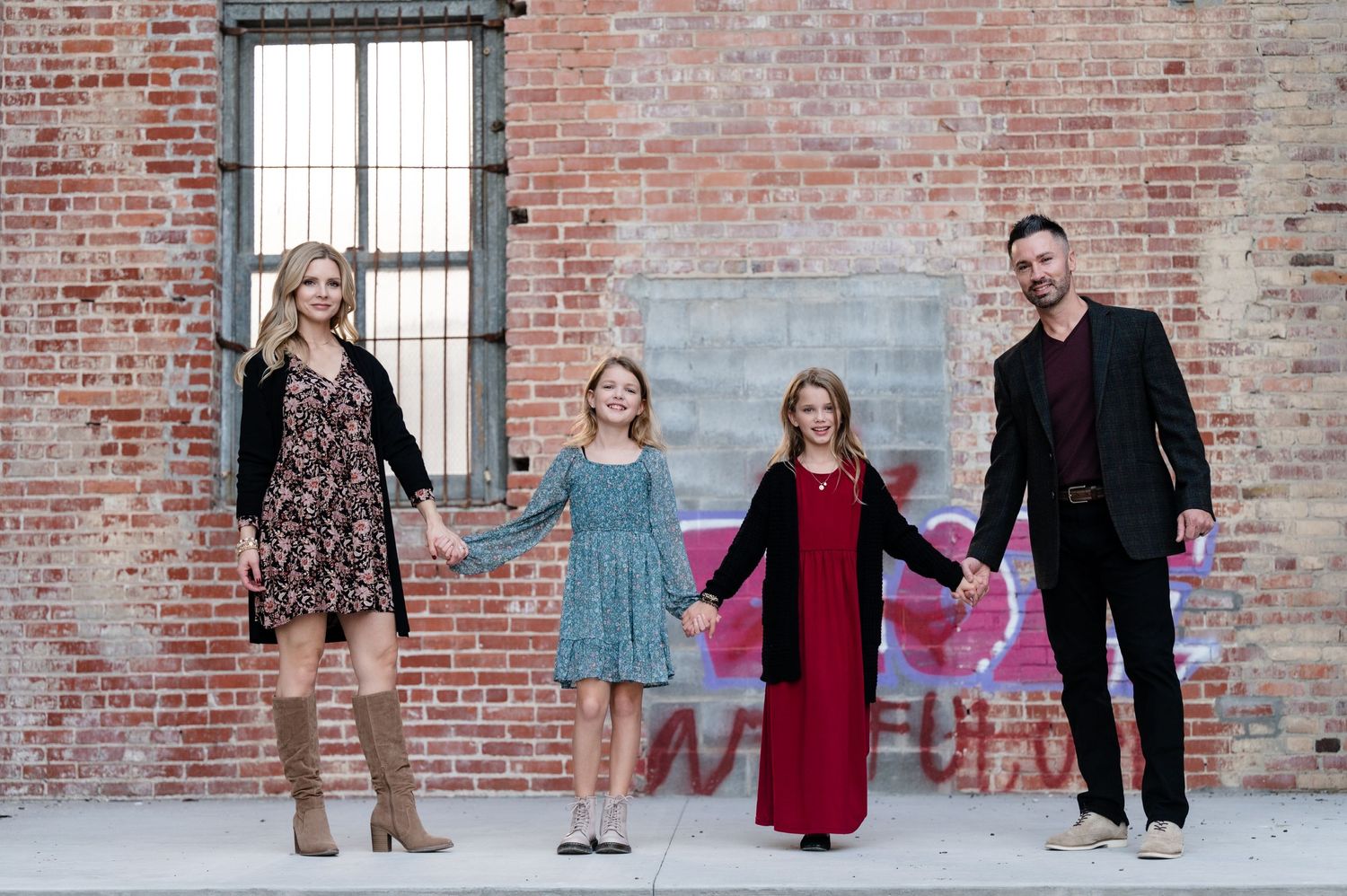 A family of four holds hands in front of an urban brick wall setting.