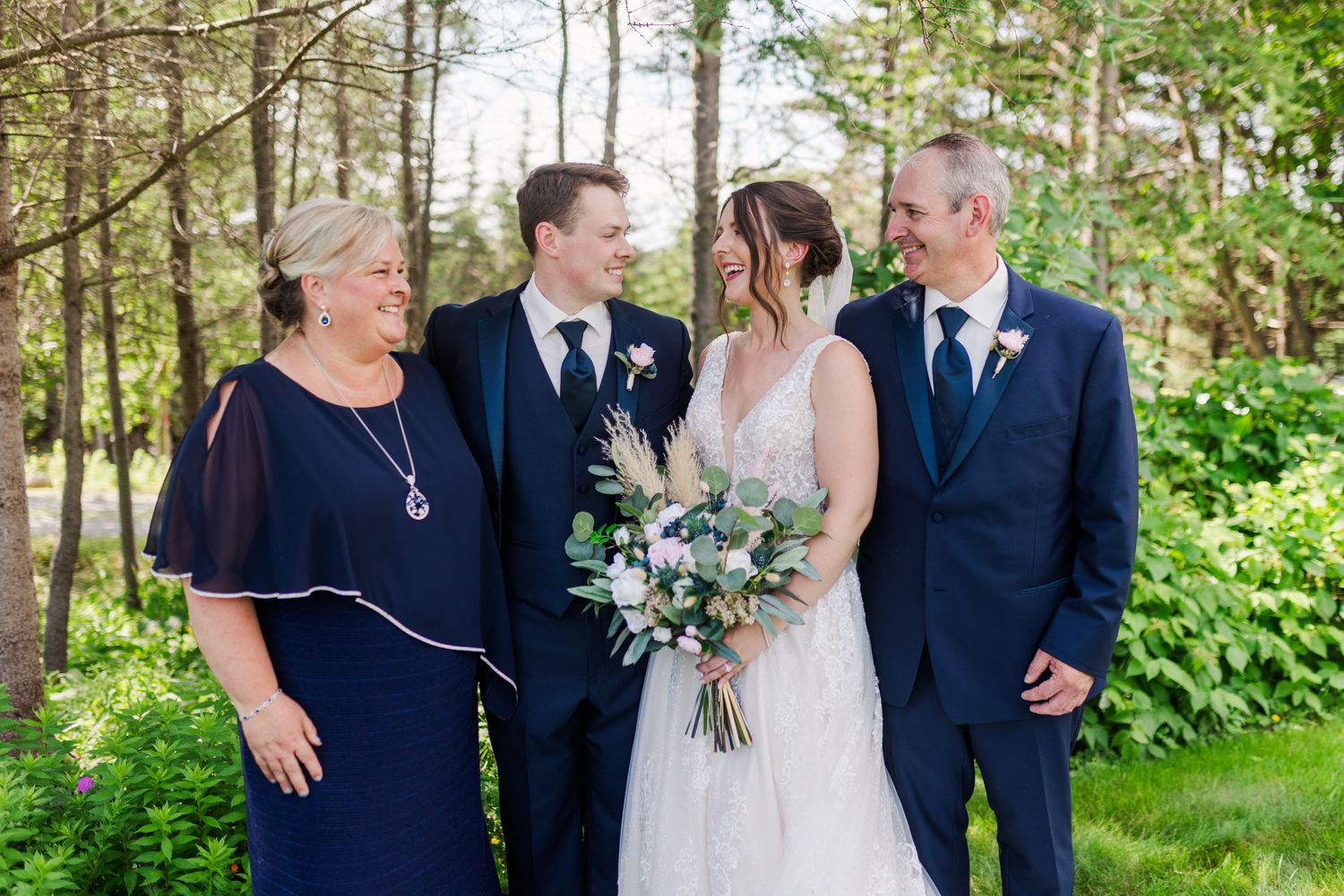 Wedding party in navy suits and white dress gather under trees for an intimate outdoor photo.