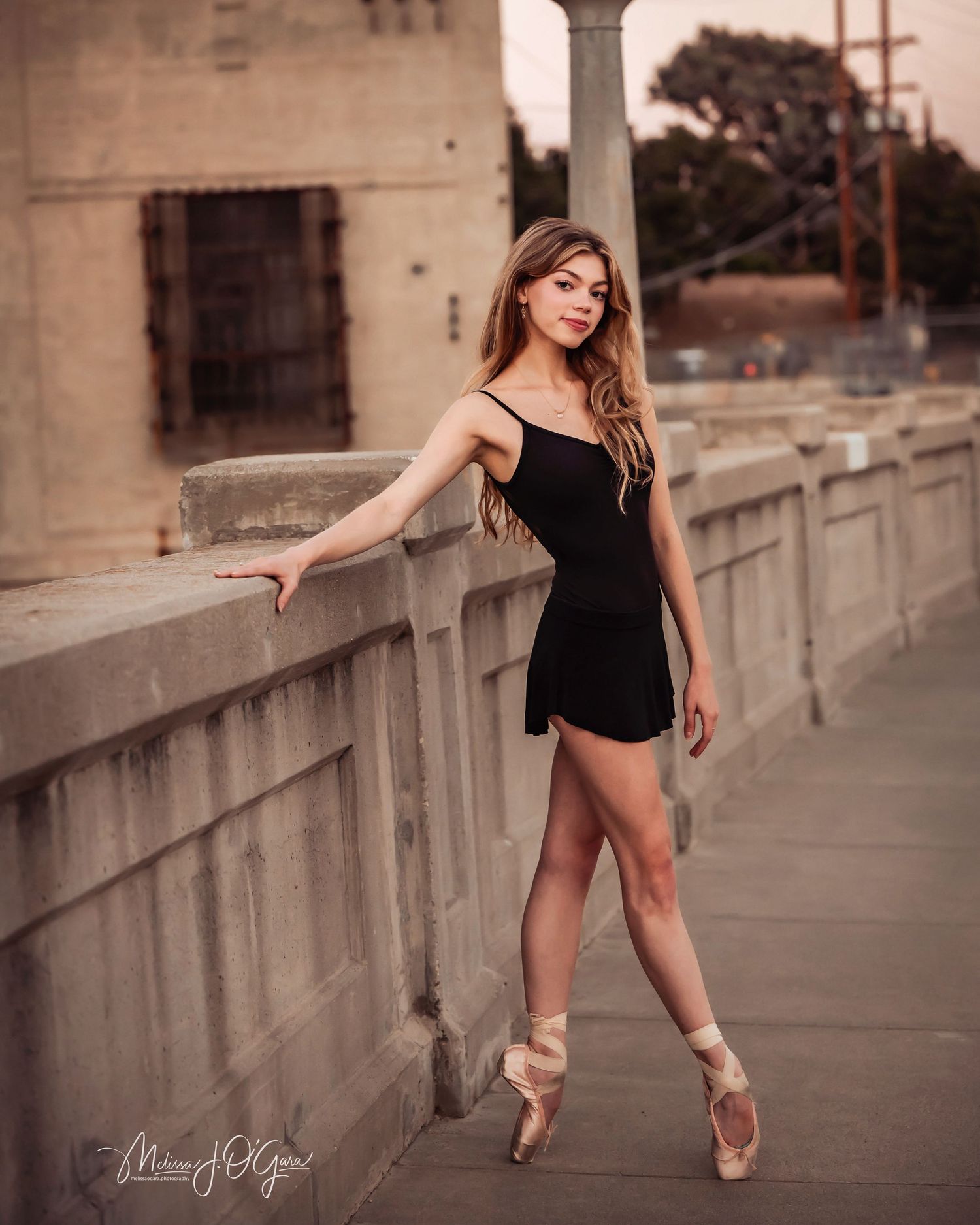 Ballet dancer in black dress poses gracefully on concrete bridge during golden hour sunset.