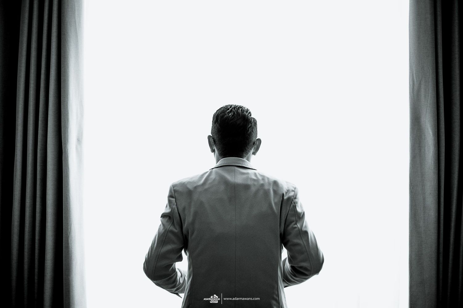A silhouetted figure in a suit stands facing curtained windows in a dramatic black and white shot.
