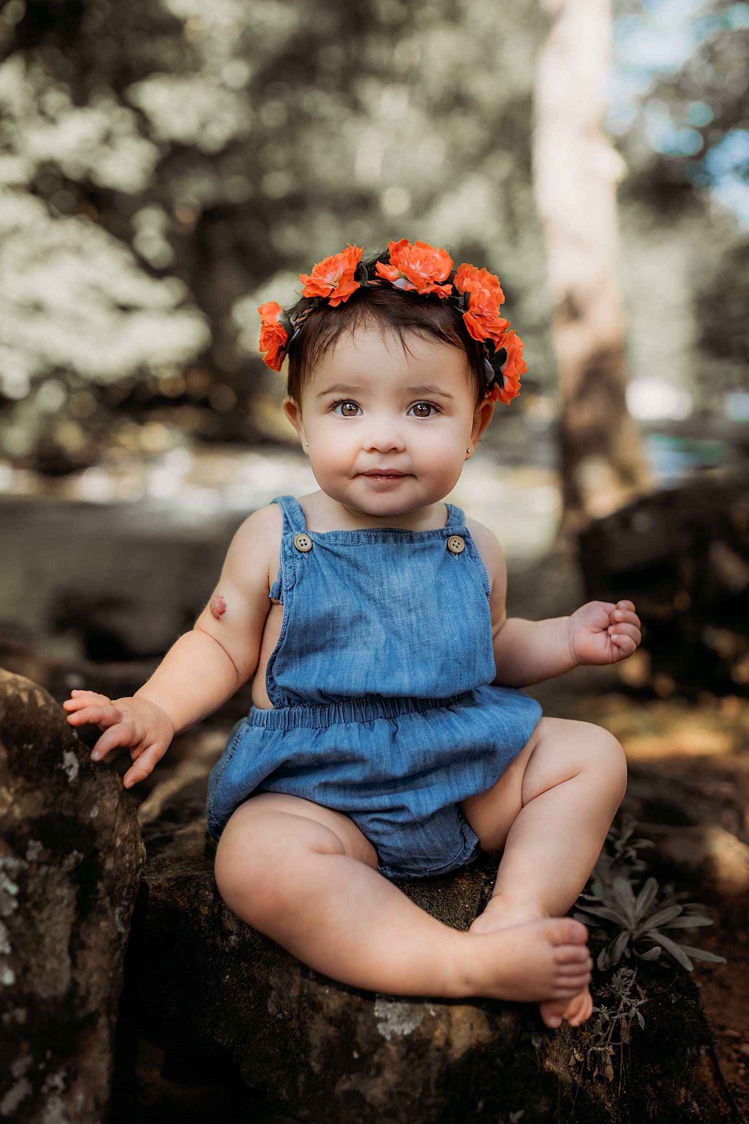 A small baby wearing a denim romper and orange flower crown sits on a rustic wooden surface in dappled sunlight.