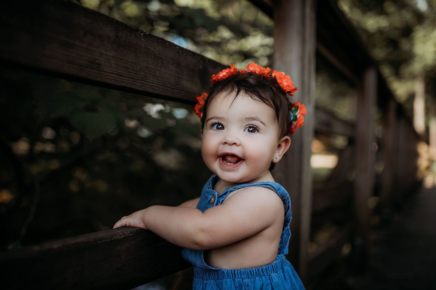 Baby in blue denim outfit with orange floral crown poses outdoors in natural lighting.