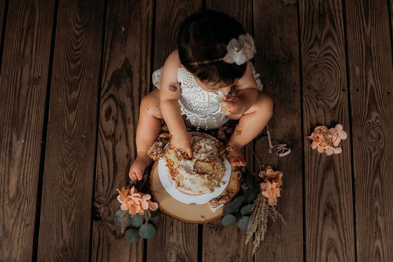 Overhead view of a rustic cake smash setup on wooden floorboards with floral decorations.