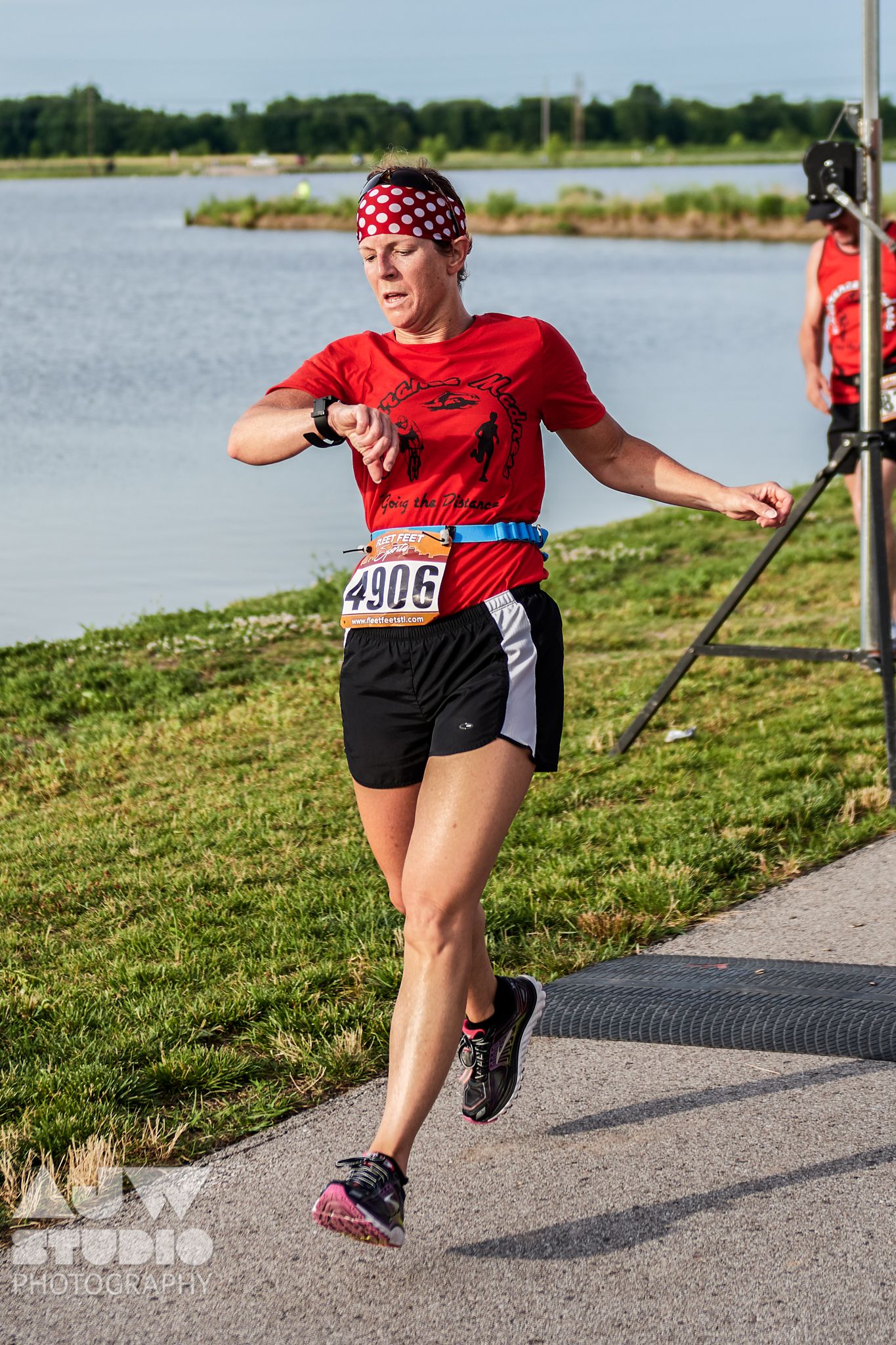 Runner in red shirt and polka dot bandana races along lakeside path during sunny competition.