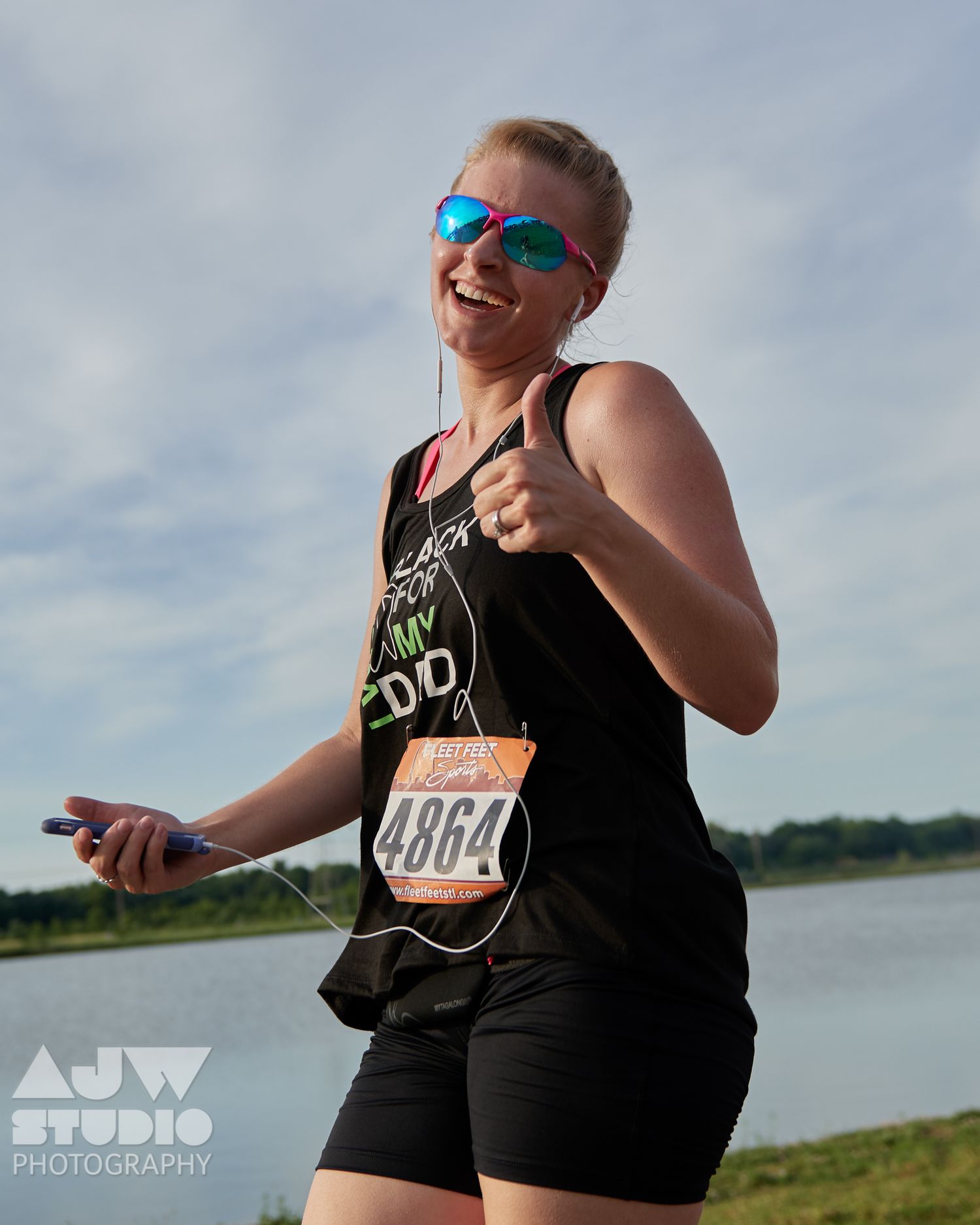 Runner wearing blue sunglasses and race bib gives thumbs up while running along waterfront path during race event.