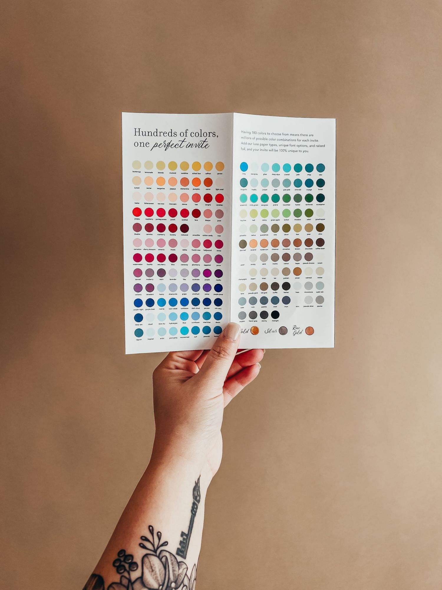 A tattooed arm holds out an open pamphlet from Basic Invite showing all of their color options for holiday card designs