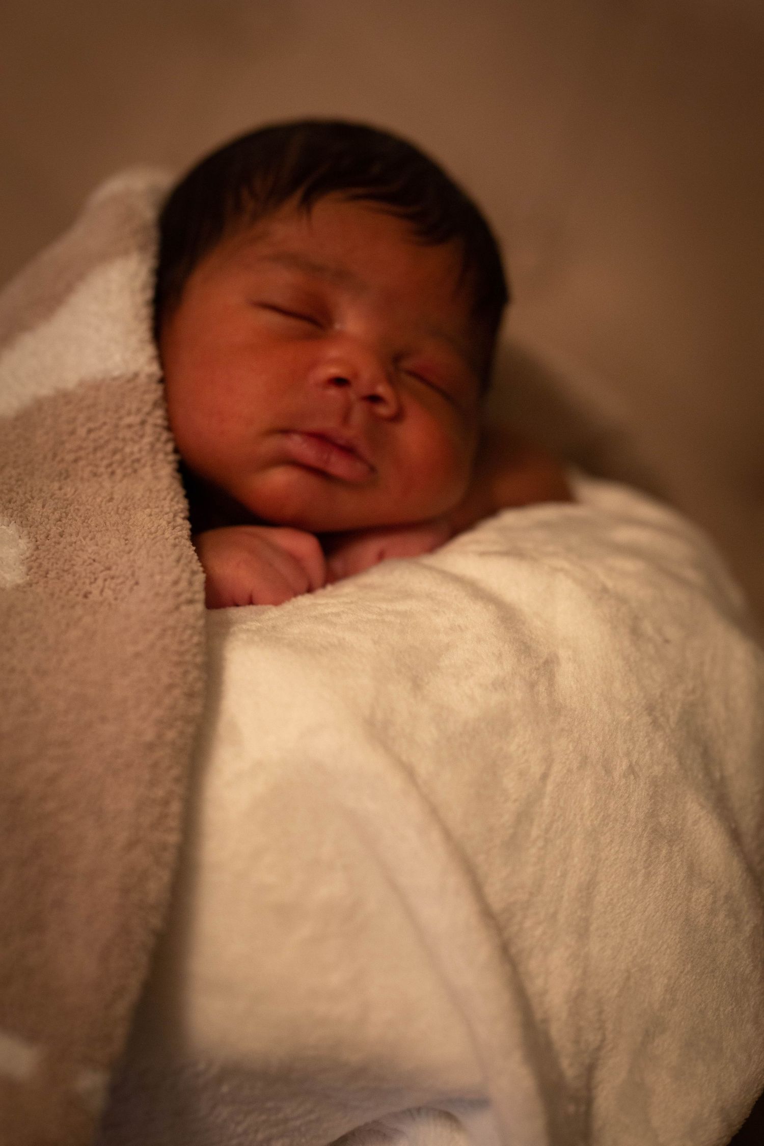 Sleeping newborn wrapped in soft white blanket in warm lighting.