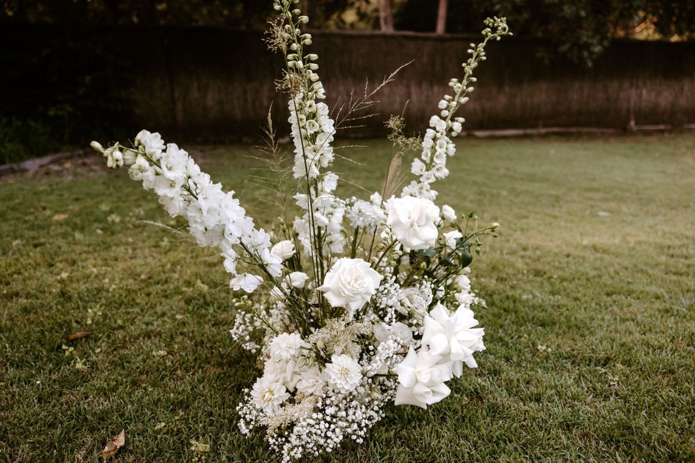 Elegant white floral arrangement with delicate blooms and greenery displayed on grass at an outdoor wedding.