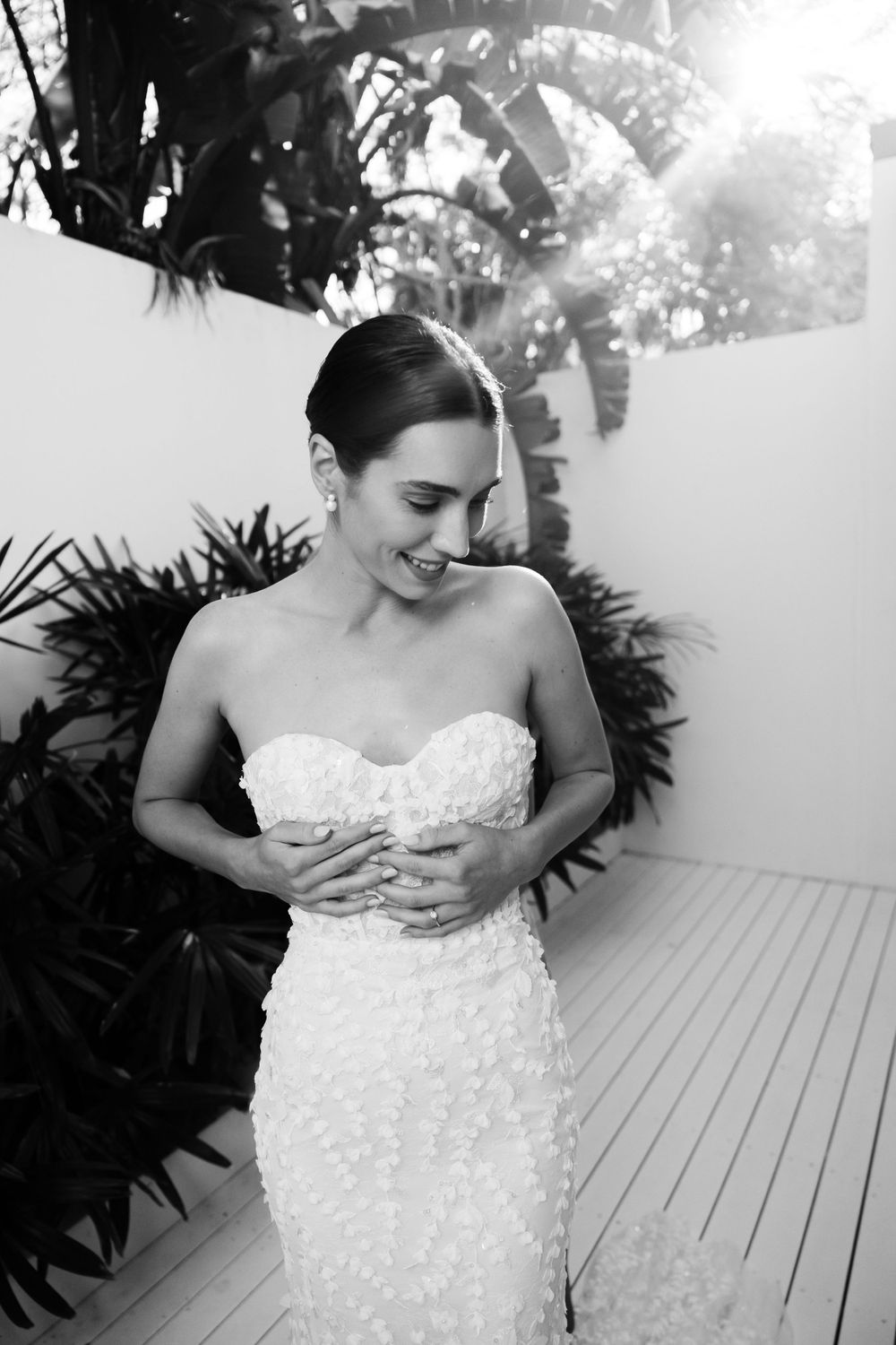 A bride in a strapless beaded wedding gown smiles in a tropical garden setting.