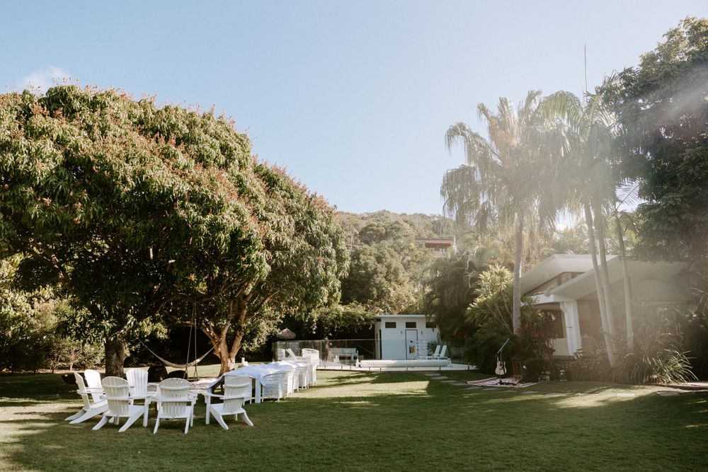 White Adirondack chairs arranged on a sunny lawn with palm trees and a white building in the background.