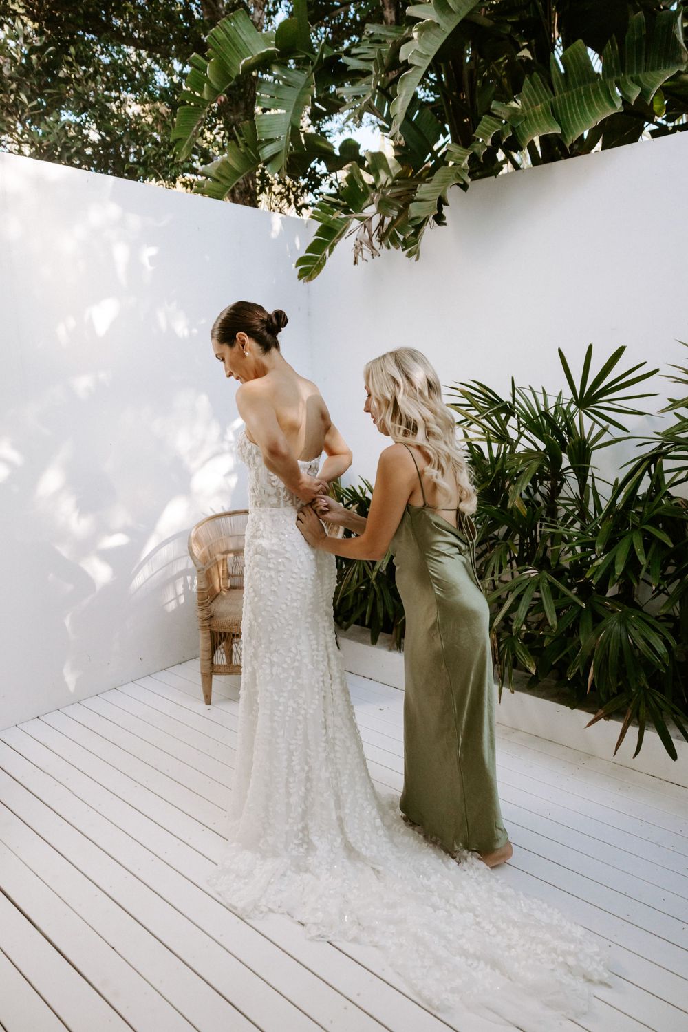 A bride getting final adjustments to her fitted lace wedding gown against a white wall with tropical plants.