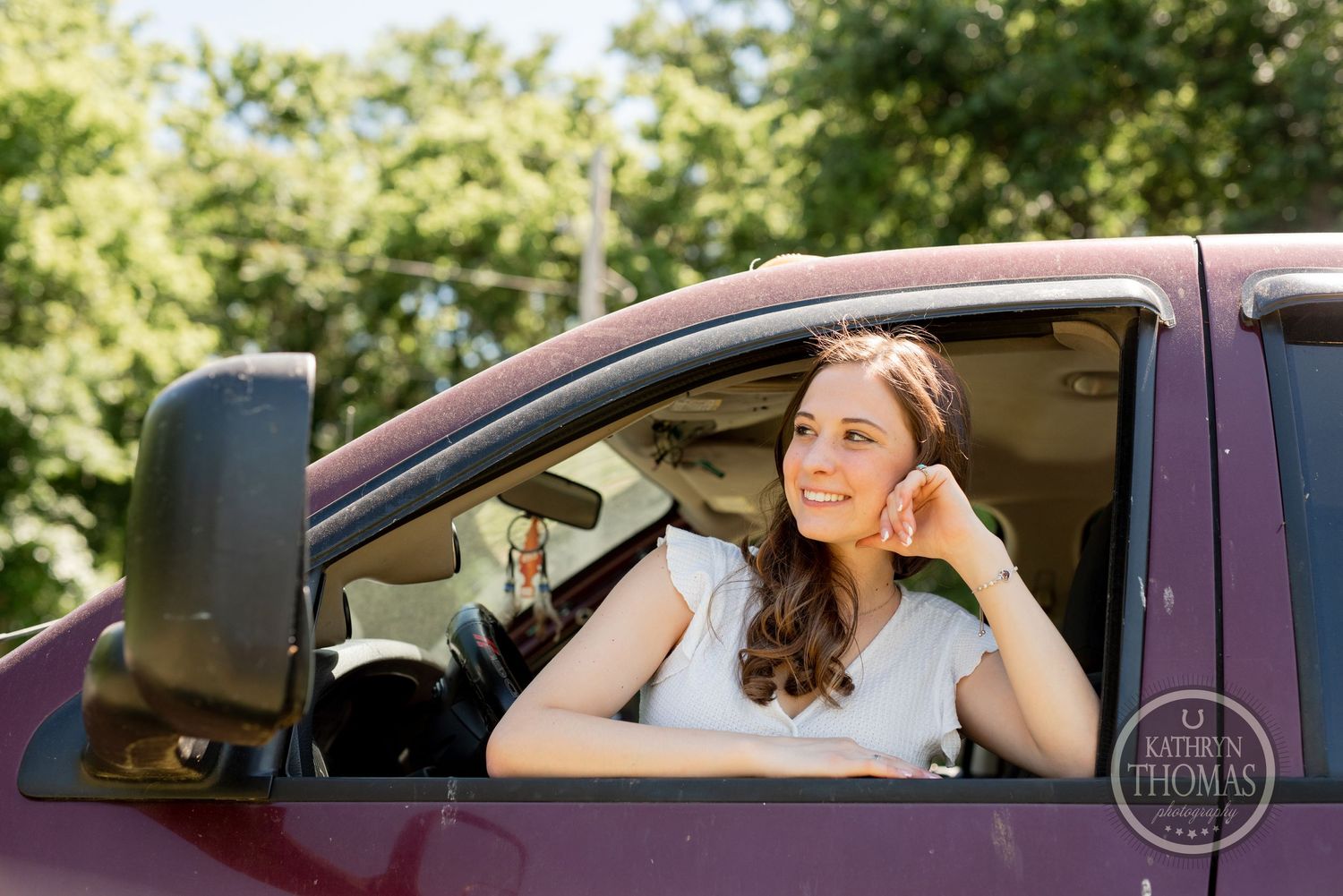 Someone poses while seated in a maroon vehicle with trees visible in the background.