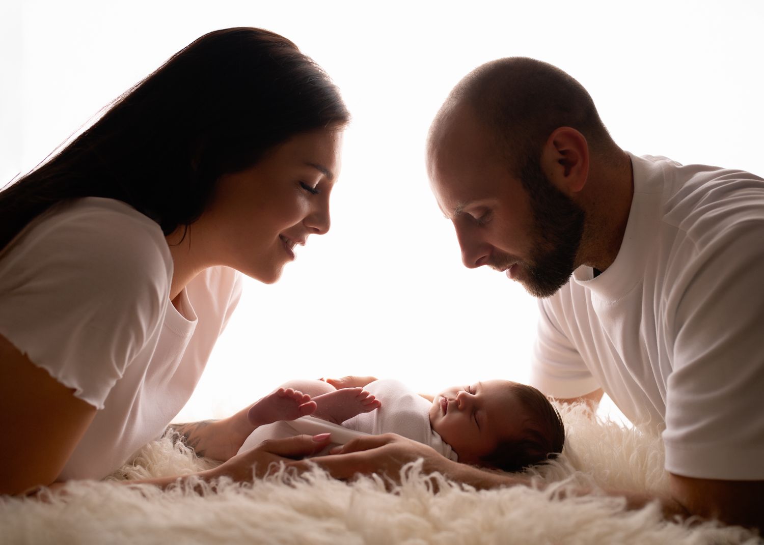 Parents gazing lovingly at newborn baby on white fur backdrop.